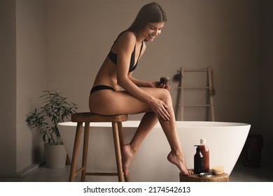 Young woman in bikini applying body peeling coffee scrub on legs at home sitting on high wooden stool against modern bathtub in luxury bathroom. Beauty and spa procedures