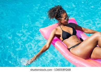 Young woman in bikini floating on inflatable mattress relaxing in the swimming pool. Beautiful fashion black girl lying on pink mat in swimming pool resort. African girl enjoying at luxury spa.
