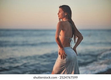 A young woman in a bikini kneels on a rocky beach at sunset. The soft evening light highlights her hair and silhouette as gentle waves wash up on the shore. Peaceful moment by the sea. - Powered by Shutterstock