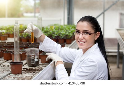 Young woman biologist in white coat holding test tube with orange chemistry in front of sprouts in flower pots - Powered by Shutterstock