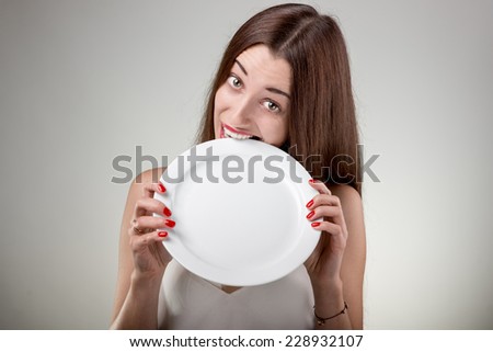 Young woman bites empty plate. Hungry woman with plate in studio on white background