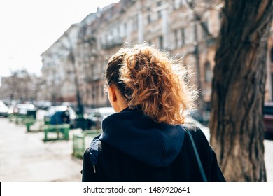 Young Woman In Black Coat And With Curly Hair Walking Through An Old European City Street Next To Road And Cars On Autumn Day, View From Behind.