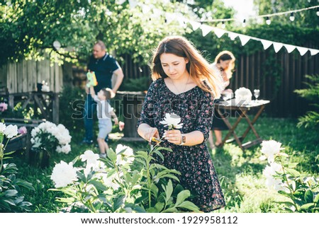 Young woman in black dress with floral pattern cuts beautiful fresh white peony flowers from bush with scissors on green summerhouse yard closeup