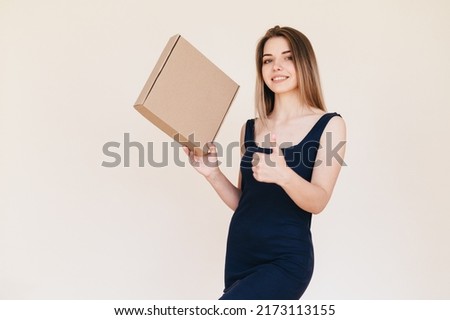 Young Woman in Black Dress Showing Thumb Up on Beige Background. Girl Holds Cardboard Box in her Hands and Smiles Broadly. Young Delivery Manager.