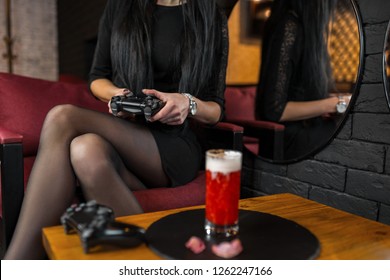 Young woman in a black dress sits in a bar with cocktail holding a black joystick in her hands and playing a modern video game. Gambling girl gamer. Interesting party.  - Powered by Shutterstock