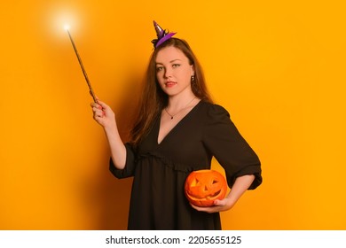 Young Woman In Black Dress And Small Purple Witch Hat Holding Orange Jack O Lantern Pumpkin And Looking At Camera. Wizard Girl Holding Luminous Magic Wand Or Glowing Magic Stick On Yellow Background.