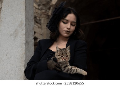 Young woman with black hair, dressed in black Gothic style, holding a cat in her lap.