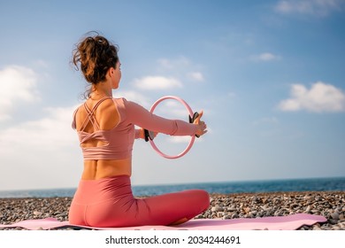 Young woman with black hair, fitness instructor in pink sports leggings and tops, doing pilates on yoga mat with magic pilates ring by the sea on the beach. Female fitness daily yoga concept. - Powered by Shutterstock