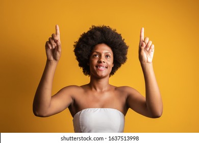 Young Woman With Black Hair Pointing To The Up With A Finger To Present A Product Or An Idea While Looking Forward Smiling On Isolated Yellow Background. Afro Woman Pointing