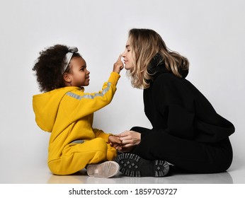 Young Woman In Black Jeans And Hoodie Sits Face To Face With Her Curly Dark-skinned Daughter Kid Girl In Yellow Sport Jumpsuit Playing Over White Wall Background. Kid Touches Her Mother Nose