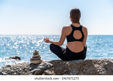 Young woman in black leggings and black top with long flowing hair practicing stretching outdoors by the sea on a sunny day. Women's yoga, fitness, pilates. The concept of a healthy lifestyle, harmony