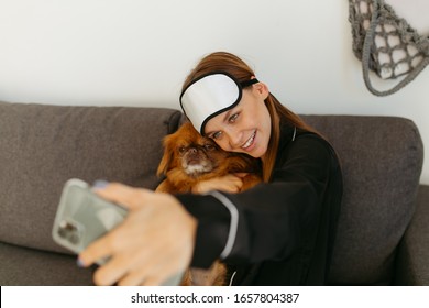 Young Woman In Black Pajamas And Bandage For Sleeping. Girl Photographs Herself With A Dog On The Phone. Grey Sofa.