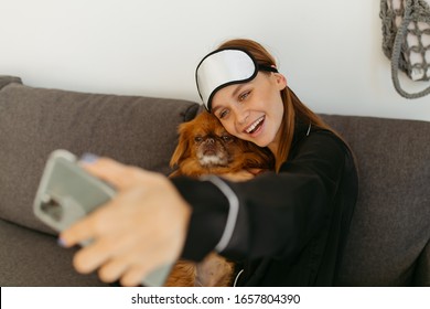 Young Woman In Black Pajamas And Bandage For Sleeping. Girl Photographs Herself With A Dog On The Phone. Grey Sofa.