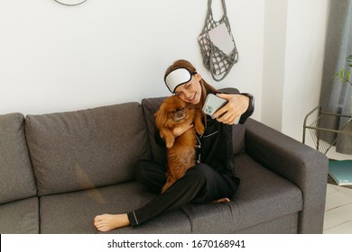 Young Woman In Black Pajamas And Bandage For Sleeping. Girl Photographs Herself With A Dog On The Phone. Grey Sofa. String Bag On A White Wall.