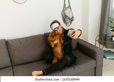 Young Woman In Black Pajamas And Bandage For Sleeping. Girl Photographs Herself With A Dog On The Phone. Grey Sofa. String Bag On A White Wall.