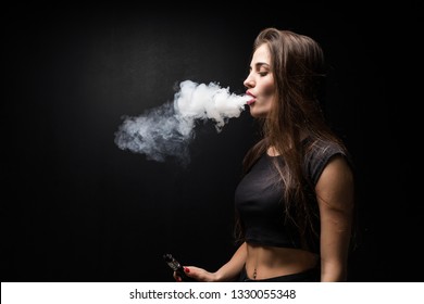 Young woman in black smokes an electronic cigarette isolated on dark background