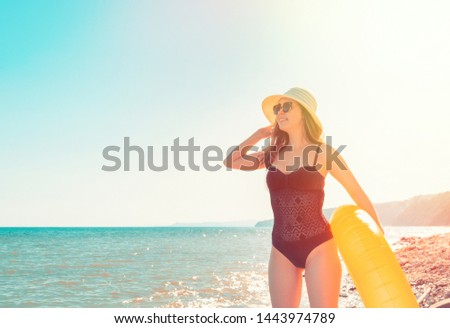 A young woman in a black swimsuit and hat is holding an orange swimming circle and enjoying the view.The sea in the background.The concept of holiday resort and travel. Copy space and tint