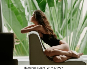 Young woman in a black swimsuit relaxing in a modern chair, gazing out large window at lush green plants - Powered by Shutterstock