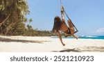 A young woman in a black swimsuit swings on a rope swing on a tropical beach, surrounded by palm trees and clear blue water. Perfect for summer, travel, beach lifestyle, and vacation promotions.