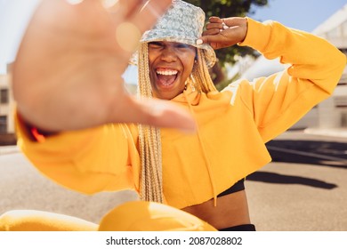 Young Woman Blocking The Camera With Her Hand. Happy Young Woman Smiling At The Camera Cheerfully While Sitting Outdoors On A Summer's Day. Vibrant Young Woman Wearing Stylish Long Braids.