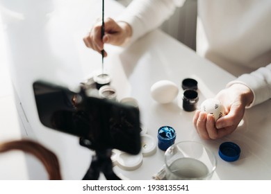 Young woman blogger live streaming online workshop. Showing how to decorate Easter eggs. Tripod with smartphone, basket with eggs, paint and laptop. Traditional holidays during COVID lockdown concept - Shutterstock ID 1938135541
