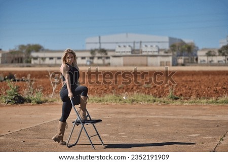 Young woman, blonde and athletic, in a black body and military boots, strong, independent and empowered, resting her foot on an outdoor chair. Concept of empowerment, strength, independence, security