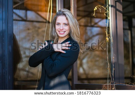 Young woman, blonde, European descent. A woman posing in a black jacket, in the background a closed street cafe. Photo taken in a winter, snowy day
