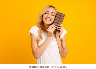 A young woman with blonde hair is shown holding a chocolate bar against a yellow background. She is smiling and looking happy, as if she is enjoying the chocolate. Her eyes are closed - Powered by Shutterstock