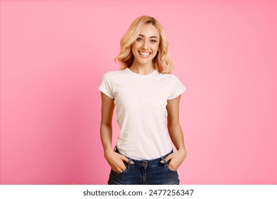 A young woman with blonde hair is smiling and looking directly at the camera. She is wearing a white t-shirt and blue jeans, and she has her hands in her pockets. Background is a bright pink color. - Powered by Shutterstock