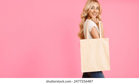 A young woman with blonde hair is smiling while holding a plain white tote bag in front of a pink background. She is wearing a white t-shirt and blue jeans. - Powered by Shutterstock