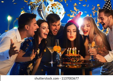 Young Woman Blowing Out Candles On Her Birthday Cake With Friends In Club