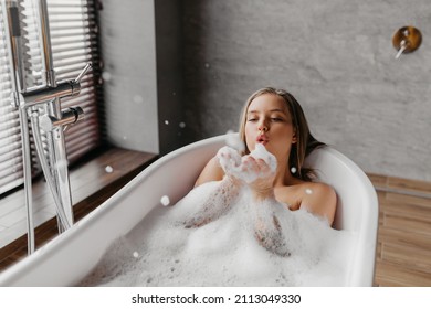 Mujer joven soplando burbujas de jabón y divirtiéndose en una bañera llena de espuma en casa, fotocopias. Dama encantadora relajándose en el baño y disfrutando de un día de belleza y cuidado de la piel