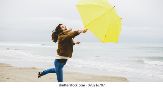 A Young Woman Is Blown Away With Her Yellow Umbrella On A Beach