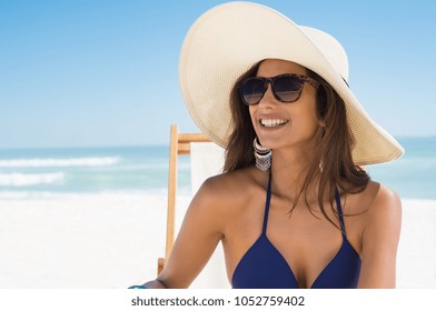 Young woman in blue bikini sitting on deck chair wearing white straw hat. Happy girl enjoying summer vacation at beach. Portrait of beautiful latin woman relaxing at beach with sunglasses.