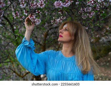 Young woman in blue dress with her hand picking flowers from a branch with white flowers in spring. Almond tree in bloom.