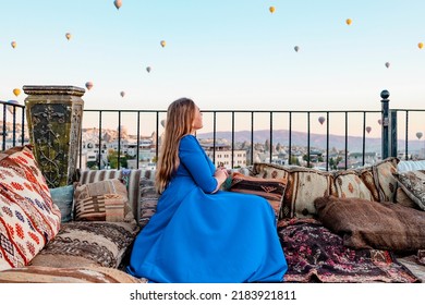 Young Woman In Blue Dress Stands On Terrace Of Hotel In Goreme Cappadocia And Looks At Hot Air Balloons Rising Into Sky, Concept  Of Must See Travel Destination, Bucket List Trip