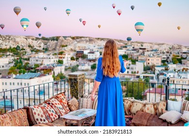 Young Woman In Blue Dress Stands On Terrace Of Hotel In Goreme Cappadocia And Looks At Hot Air Balloons Rising Into Sky, Concept  Of Must See Travel Destination, Bucket List Trip