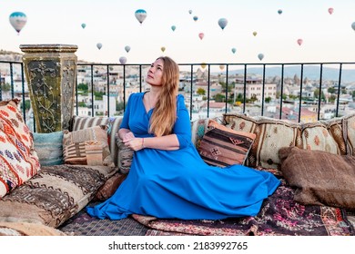 Young Woman In Blue Dress Stands On Terrace Of Hotel In Goreme Cappadocia And Looks At Hot Air Balloons Rising Into Sky, Concept  Of Must See Travel Destination, Bucket List Trip