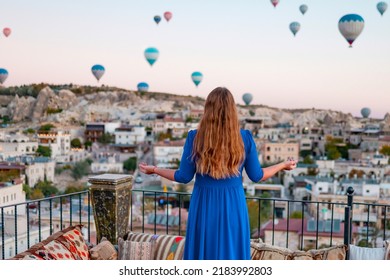 Young Woman In Blue Dress Stands On Terrace Of Hotel In Goreme Cappadocia And Looks At Hot Air Balloons Rising Into Sky, Concept  Of Must See Travel Destination, Bucket List Trip