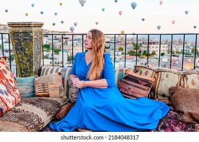 Young Woman In Blue Dress Stands On Terrace Of Hotel In Goreme Cappadocia And Looks At Hot Air Balloons Rising Into Sky, Concept  Of Must See Travel Destination, Bucket List Trip