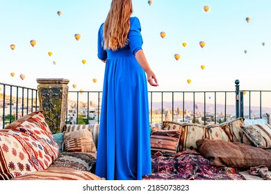 Young Woman In Blue Dress Stands On Terrace Of Hotel In Goreme Cappadocia And Looks At Hot Air Balloons Rising Into Sky, Concept  Of Must See Travel Destination, Bucket List Trip