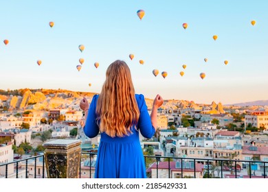 Young Woman In Blue Dress Stands On Terrace Of Hotel In Goreme Cappadocia And Looks At Hot Air Balloons Rising Into Sky, Concept  Of Must See Travel Destination, Bucket List Trip