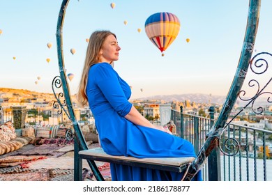 Young Woman In Blue Dress Stands On Terrace Of Hotel In Goreme Cappadocia And Looks At Hot Air Balloons Rising Into Sky, Concept  Of Must See Travel Destination, Bucket List Trip