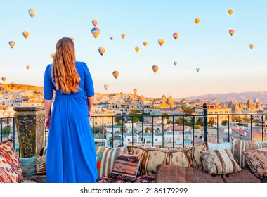 Young Woman In Blue Dress Stands On Terrace Of Hotel In Goreme Cappadocia And Looks At Hot Air Balloons Rising Into Sky, Concept  Of Must See Travel Destination, Bucket List Trip