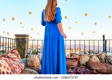 Young Woman In Blue Dress Stands On Terrace Of Hotel In Goreme Cappadocia And Looks At Hot Air Balloons Rising Into Sky, Concept  Of Must See Travel Destination, Bucket List Trip