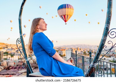 Young Woman In Blue Dress Stands On Terrace Of Hotel In Goreme Cappadocia And Looks At Hot Air Balloons Rising Into Sky, Concept  Of Must See Travel Destination, Bucket List Trip