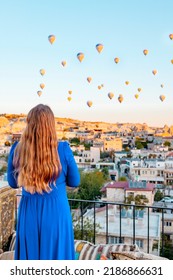 Young Woman In Blue Dress Stands On Terrace Of Hotel In Goreme Cappadocia And Looks At Hot Air Balloons Rising Into Sky, Concept  Of Must See Travel Destination, Bucket List Trip