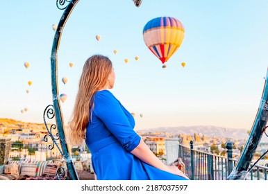 Young Woman In Blue Dress Stands On Terrace Of Hotel In Goreme Cappadocia And Looks At Hot Air Balloons Rising Into Sky, Concept  Of Must See Travel Destination, Bucket List Trip