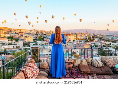 Young Woman In Blue Dress Stands On Terrace Of Hotel In Goreme Cappadocia And Looks At Hot Air Balloons Rising Into Sky, Concept  Of Must See Travel Destination, Bucket List Trip