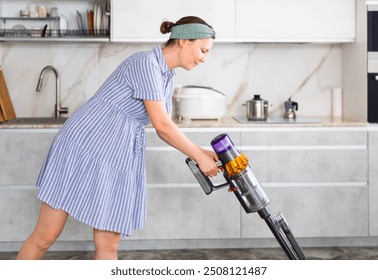 Young woman in blue dress and turban headband cleaning kitchen. Smiling housewife run vacuum cleaner floor covering near dining table - Powered by Shutterstock